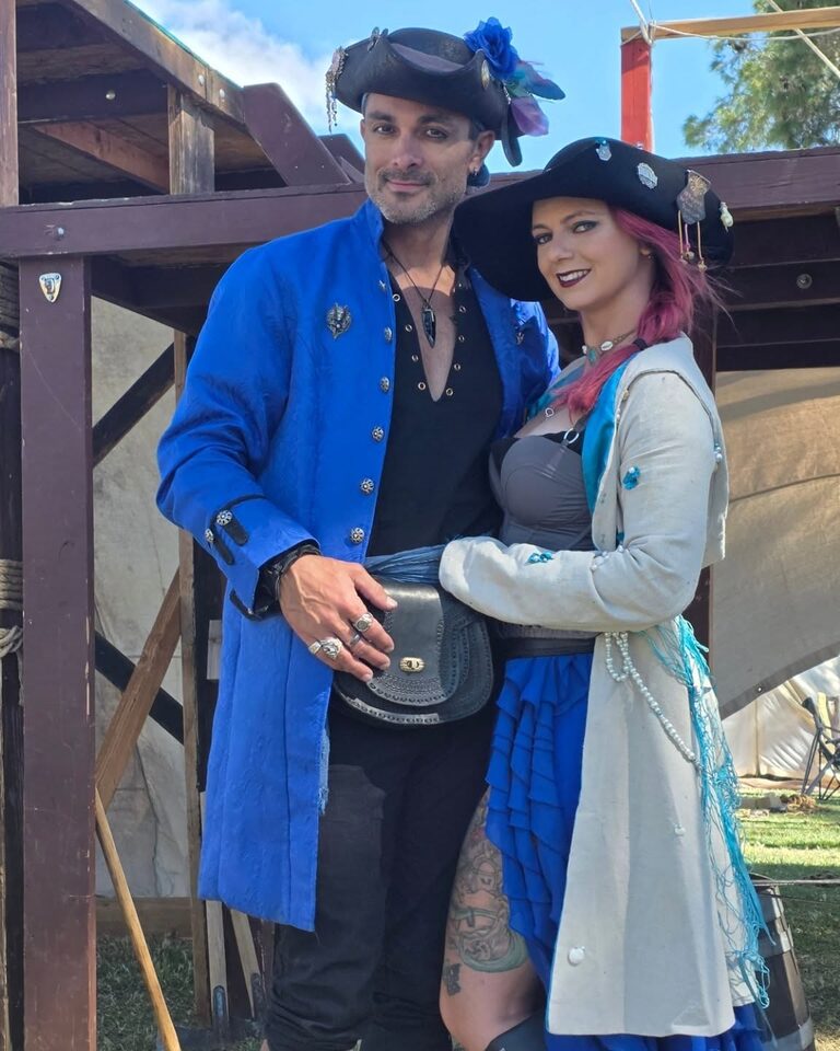 Sir Edwin in a royal blue coat and Siren KP in a pale coat, both in tricorn hats and pirate dress, standing arm in arm.