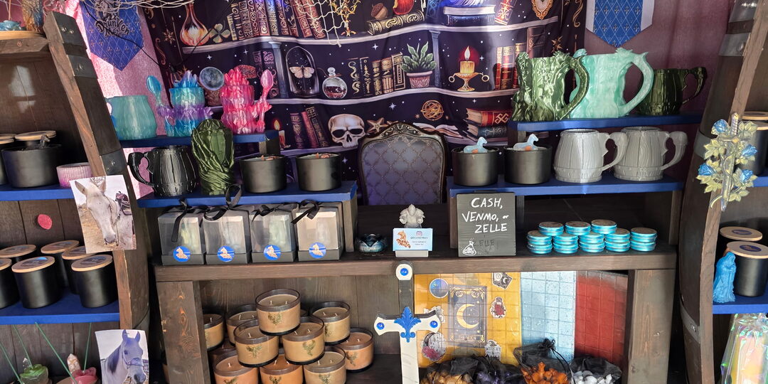 Inside the Drunken Mare stall: dark wood barrel shelves edged in royal blue, black and amber jar candles, stacked teal balm tins, a wizard-library tapestry and a fishing net strung with lights overhead.
