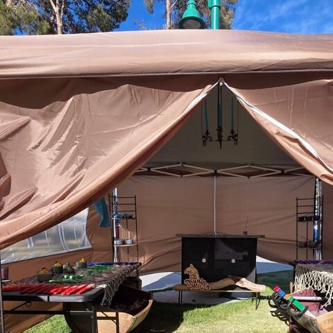 The Drunken Mare tent set up on the grass at a faire, its front flaps tied open on tables of candles.