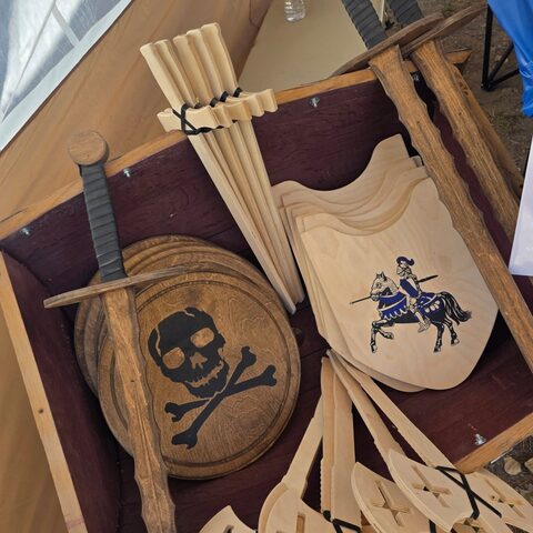Wooden pieces at our stall: practice swords, a shield painted with a knight on horseback, and a round skull-and-crossbones shield.