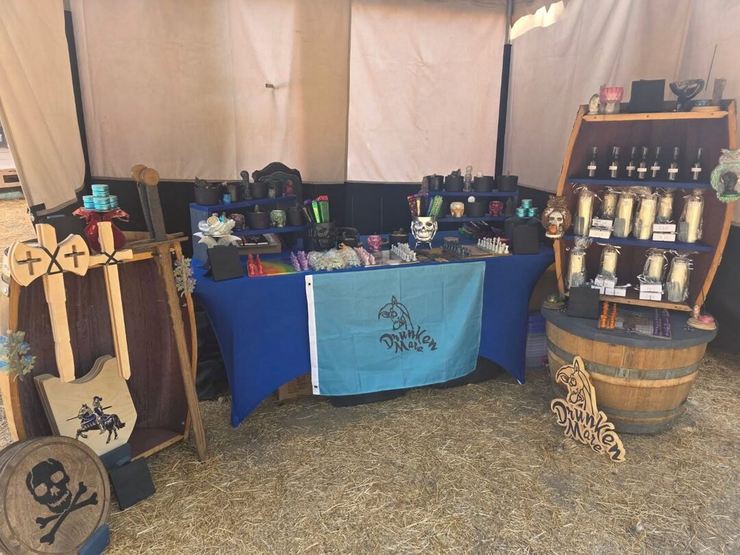 The Drunken Mare stall set up at a faire: a royal blue table of candles, skulls and chess sets under a teal Drunken Mare flag, a barrel shelf of tall candles, wooden swords and shields, and a cut-out Drunken Mare sign on the straw.