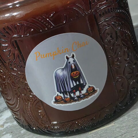 A dark embossed-glass candle jar with a round Pumpkin Chai label: a horse in a ghost sheet holding a jack-o'-lantern.